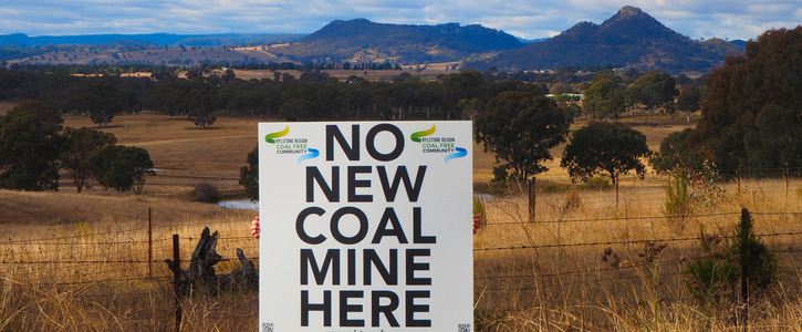 A sign reads "No new coal mine here", hung up on a barbed wire fence in front of a field of yellow grass, with green and blue hills silhouetted in the background. 